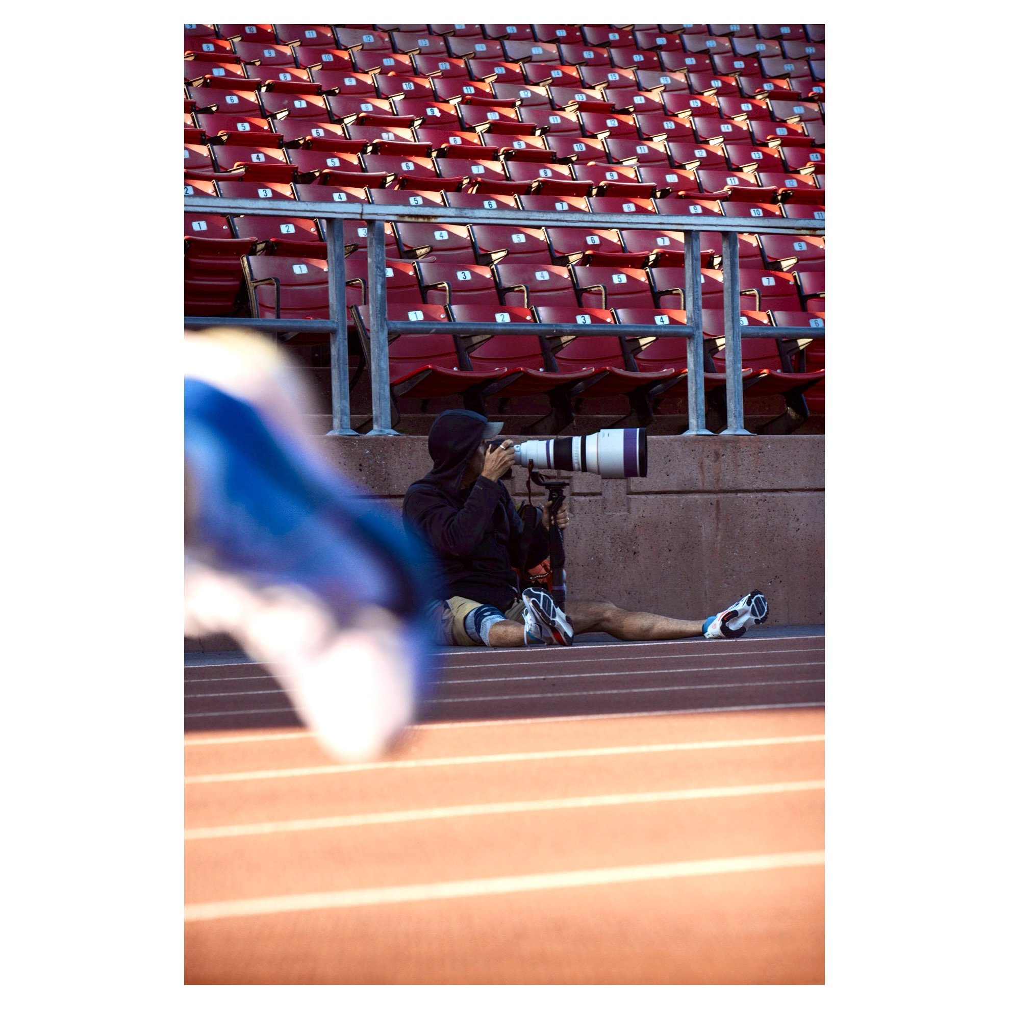 Empty red stadium seats with numbered stickers, and a photographer sitting on the ground with a large telephoto camera lens.