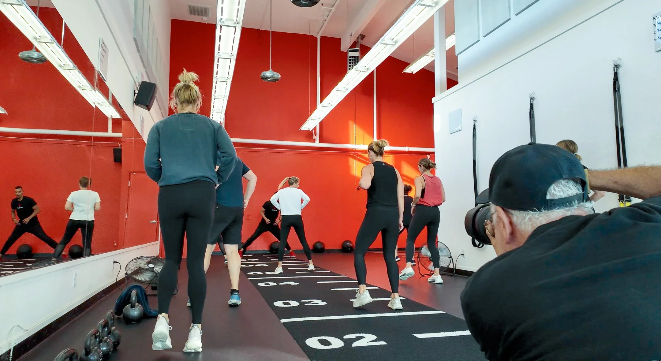 People exercising in a fitness class with a photographer capturing the scene, inside a gym with red and white walls and fitness equipment on the floor.