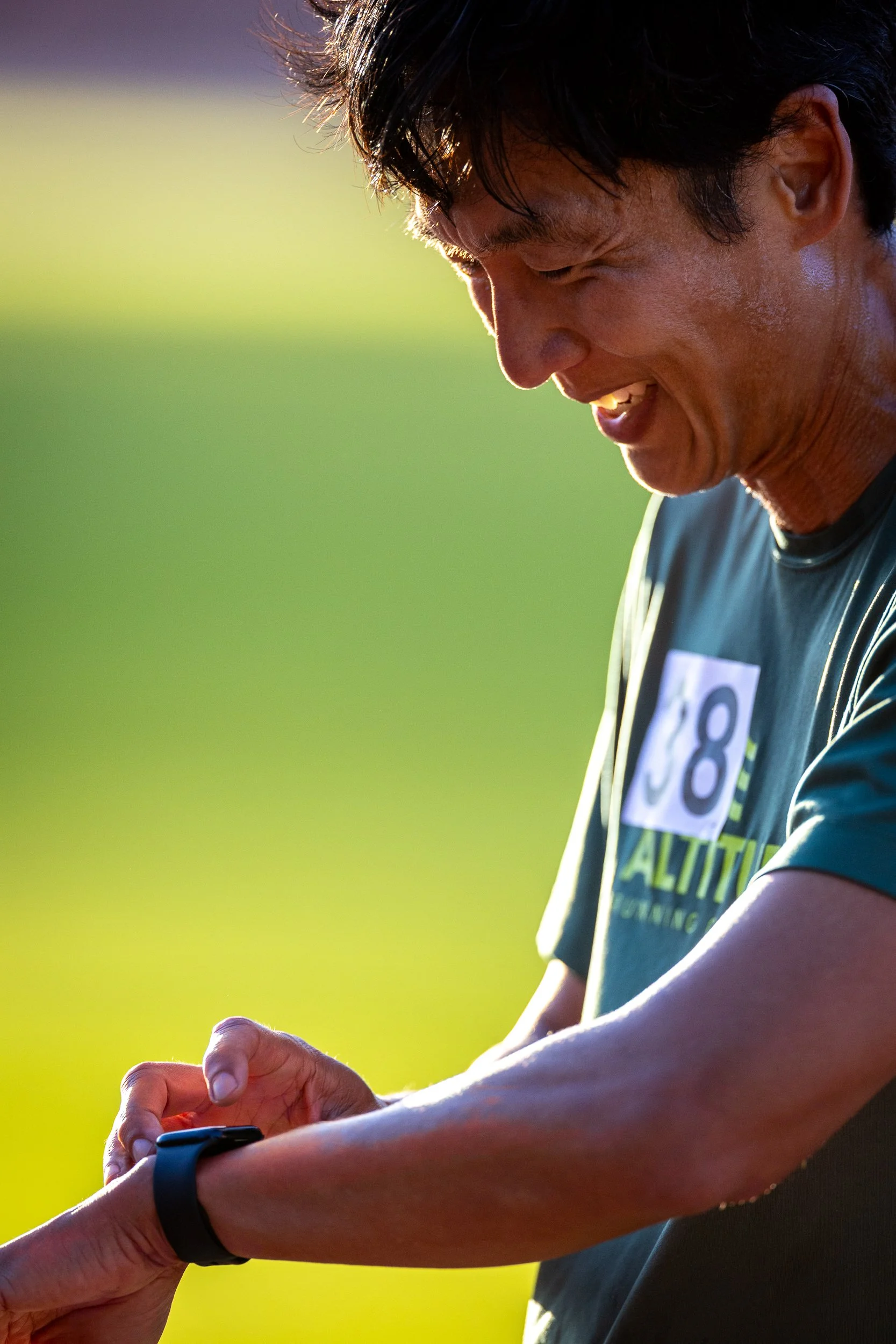A man with dark hair, sweating and smiling, wearing a black t-shirt with a bib number, looking at a smartwatch on his wrist.