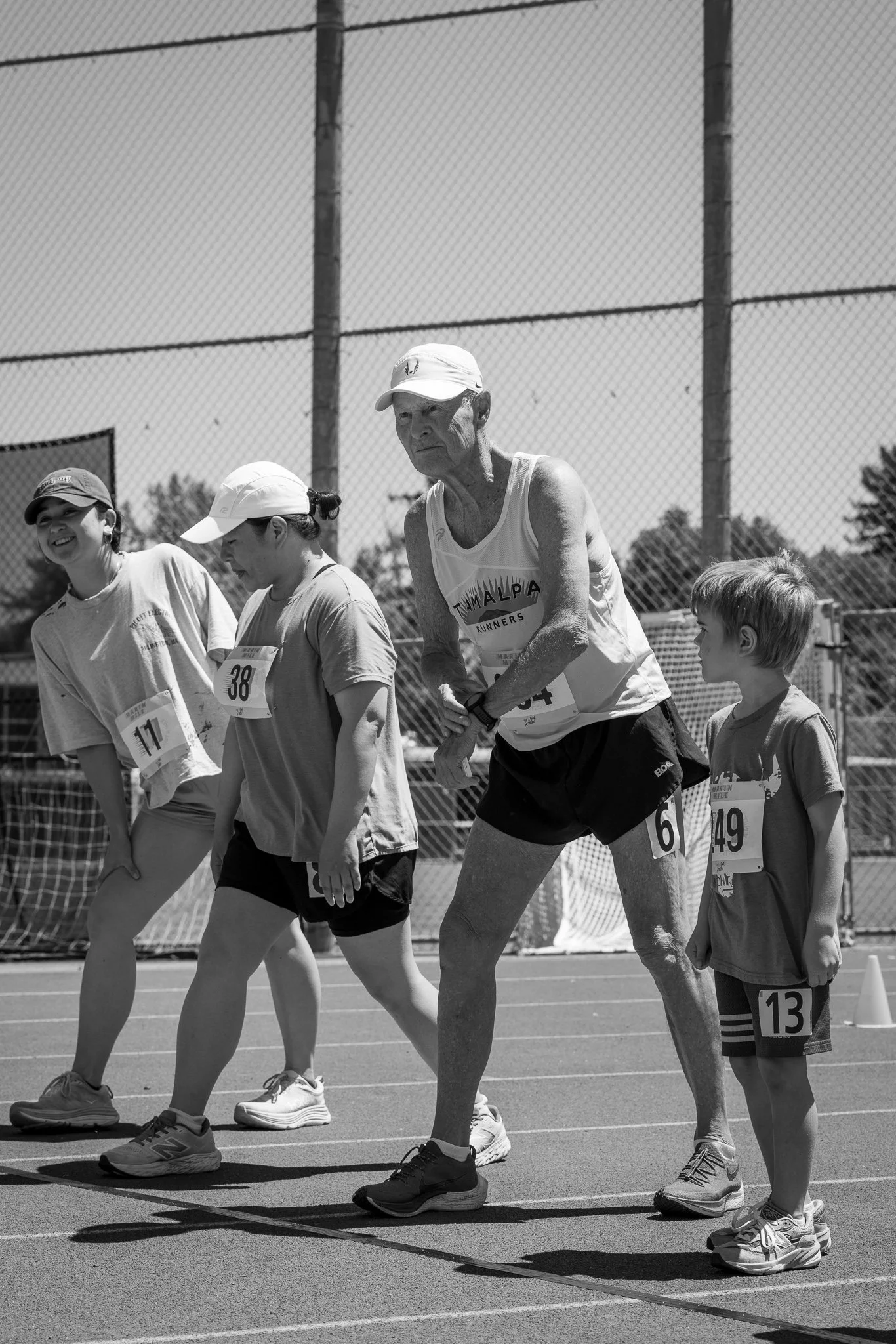 People at the starting line of a race on an outdoor track, preparing to run, with a chain-link fence and trees in the background, black and white photograph.