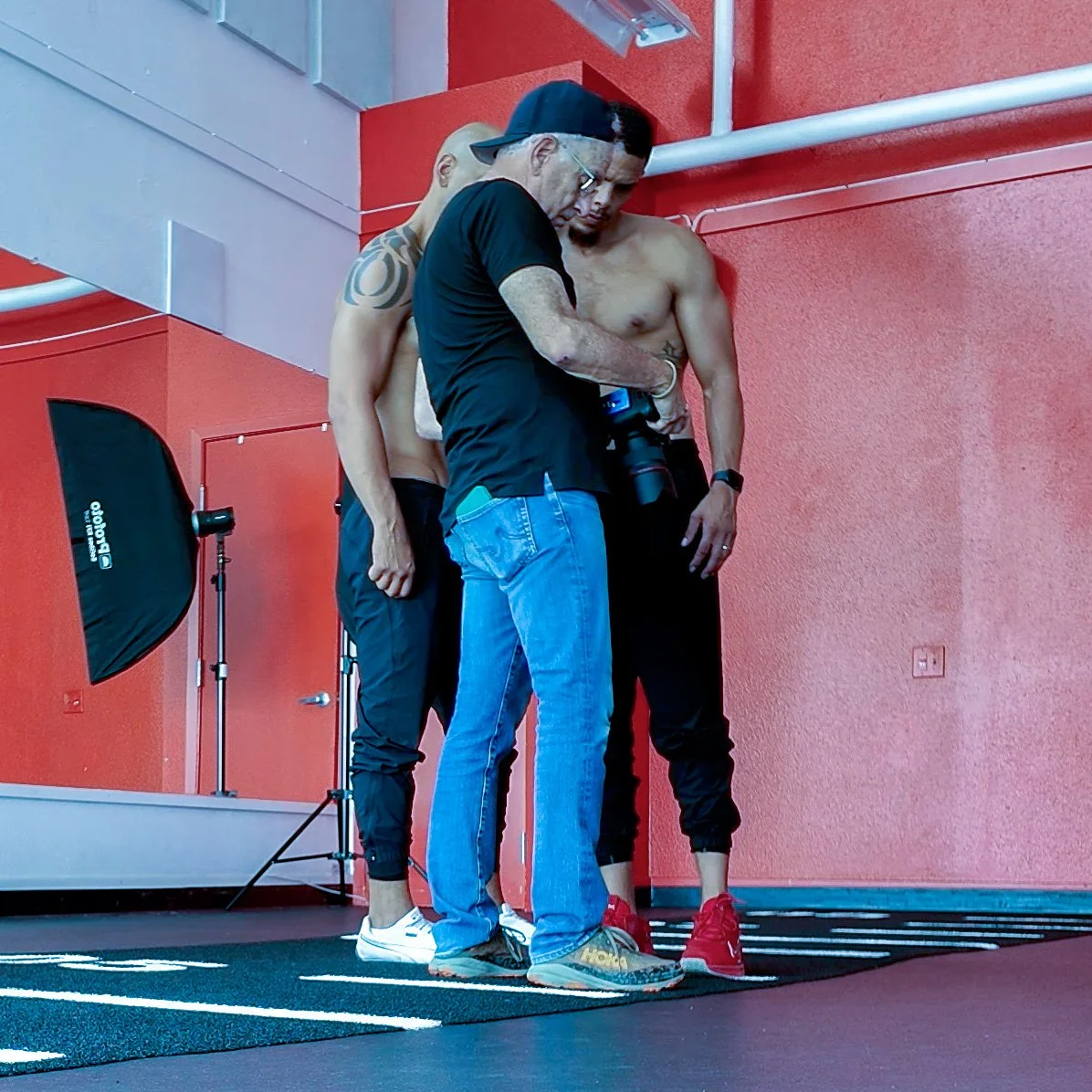 Three men standing together in a gym, closely examining something. Two are shirtless and the third wears a black shirt, cap, and jeans. The background includes a red wall, a professional light, and gym equipment.