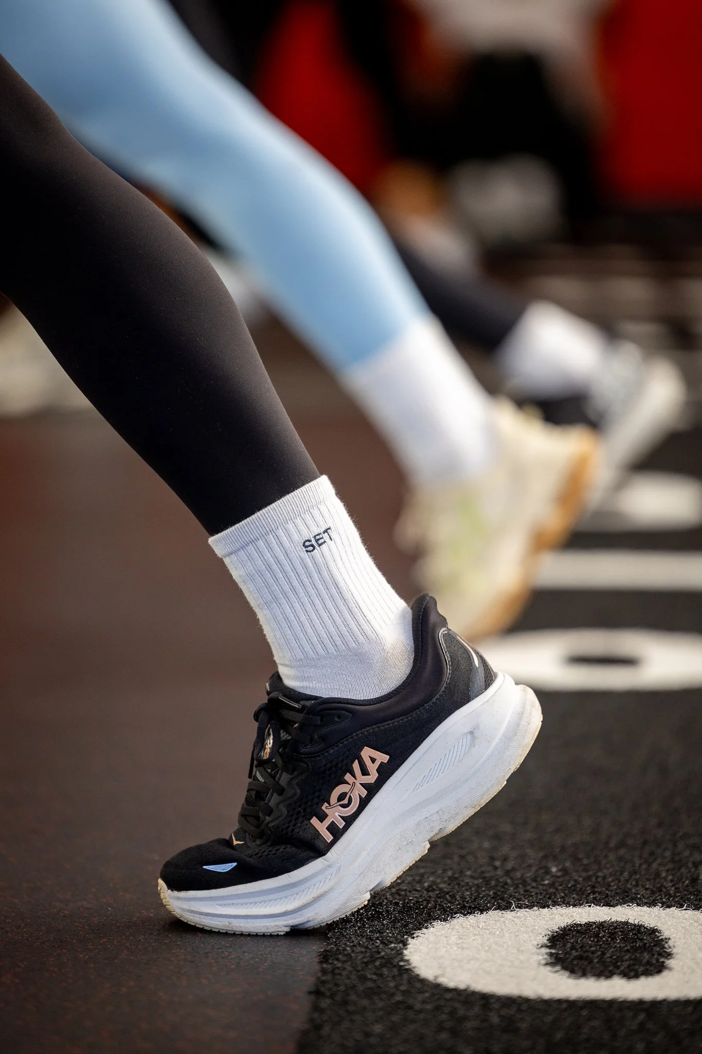 Close-up of a person wearing black athletic shoes with a white sole and black laces, white socks with the word 'SET' on the side, black leggings, and light blue workout pants. The person is on a black and white gym floor with the number 8 visible.