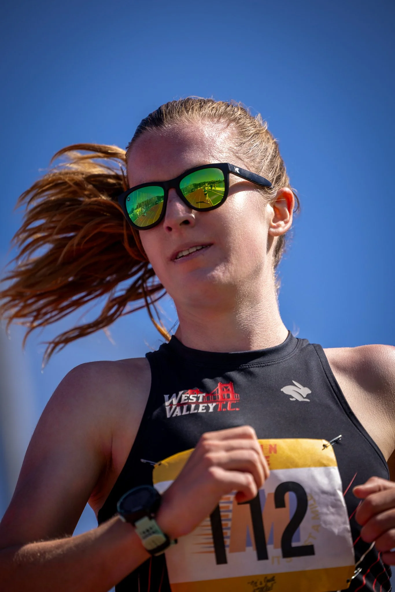 A female runner with long hair, wearing sunglasses and a black athletic jersey with a bib number 112, participating in a race under a clear blue sky.