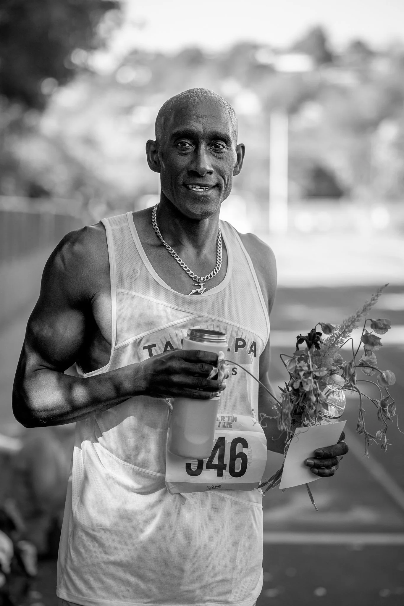 A male marathon runner holding a disposable coffee cup, a bouquet of flowers, and a piece of paper, wearing a race bib number 346, smiling at the camera.