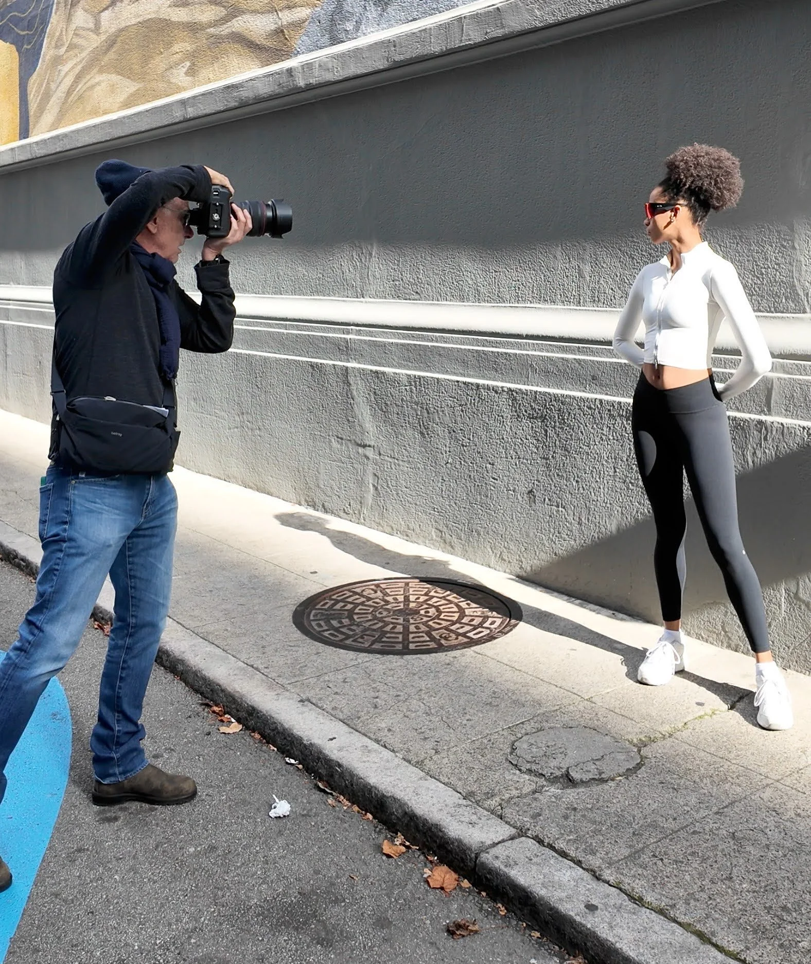 Photographer taking a picture of a woman posing on the sidewalk beside a gray wall.