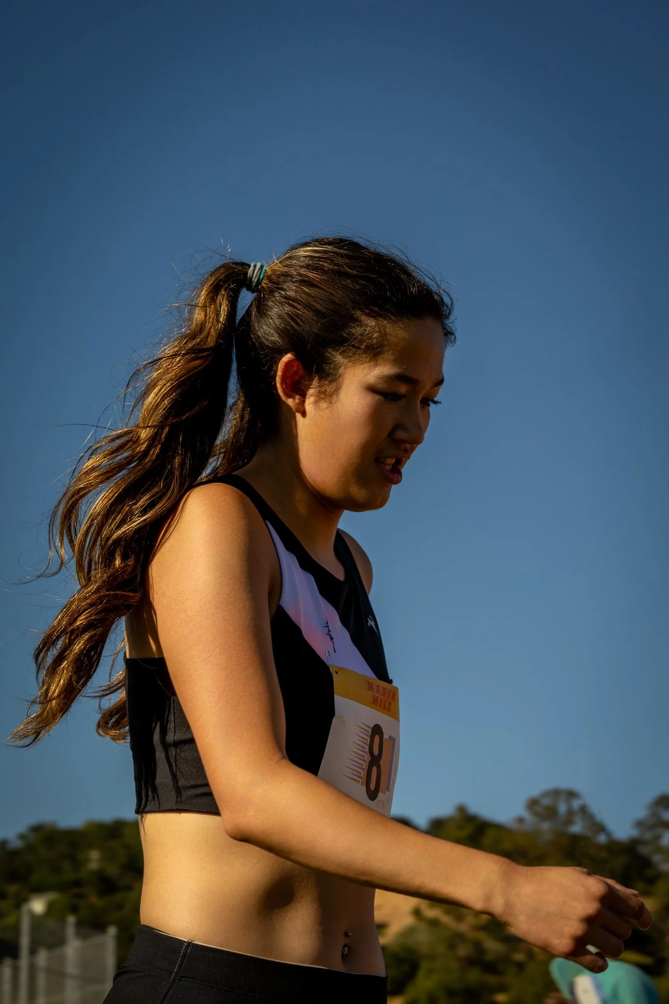 A young woman with a ponytail running in a race, wearing a black and white sports top with the number 8 bib, outdoors on a sunny day.
