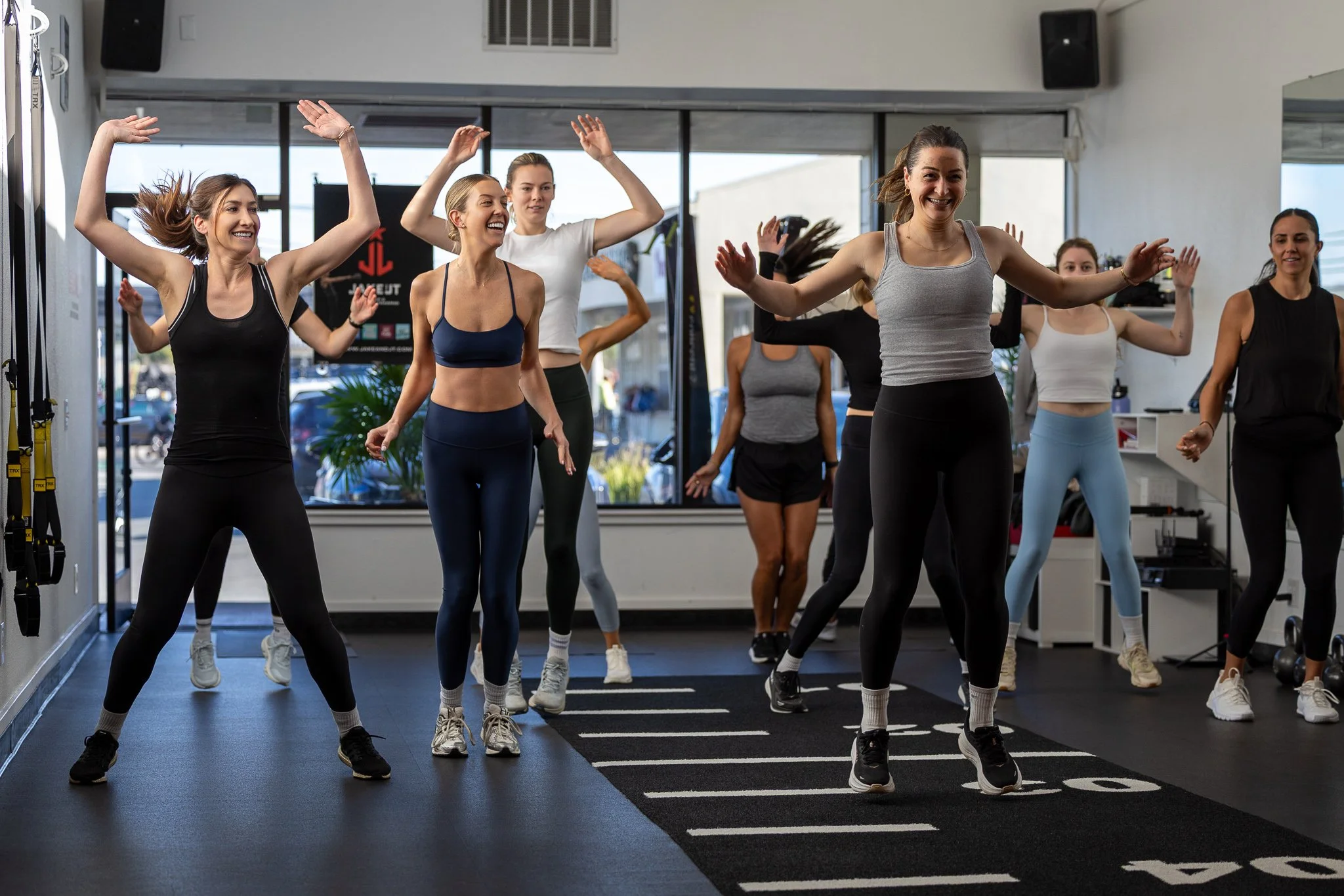 Group of women participating in a fitness class in a gym, performing aerobic exercises on a black mat with white markings.