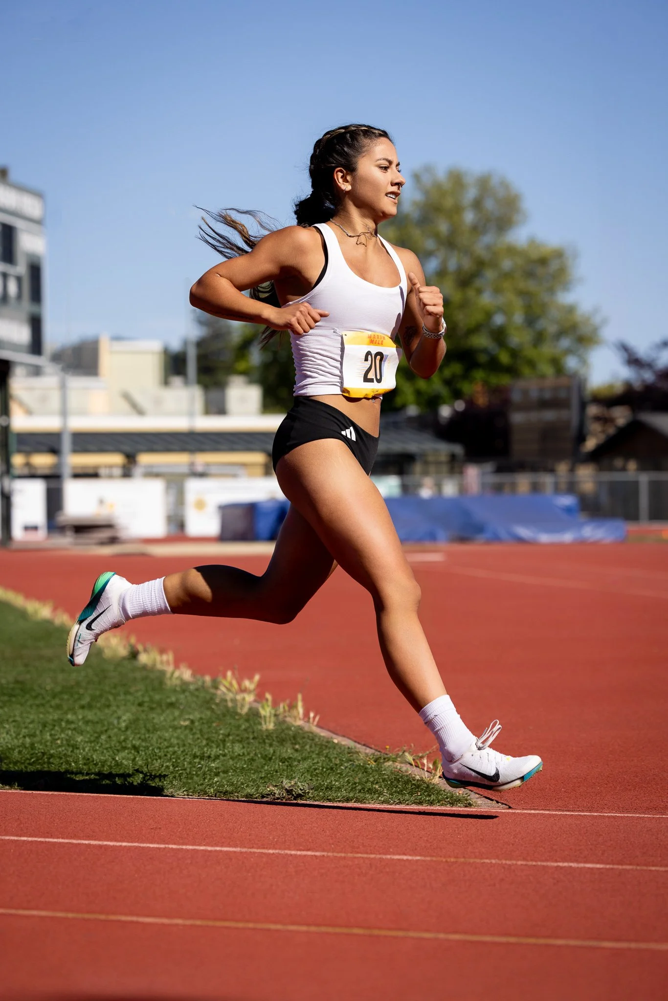 A female athlete running on a track, wearing a white sports bra, black shorts, white socks, and white running shoes.