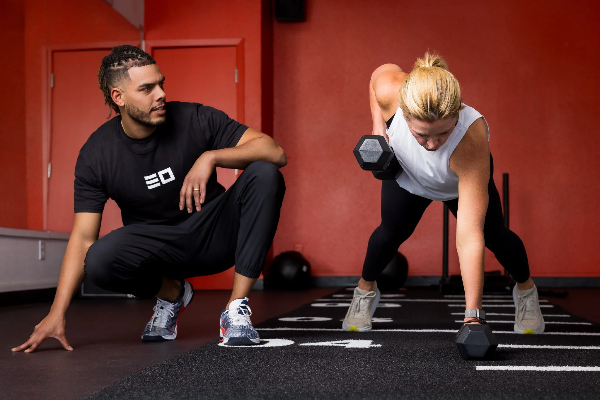 A woman is performing a plank exercise with a dumbbell in her right hand while a trainer observes her in a gym with red walls and black flooring.