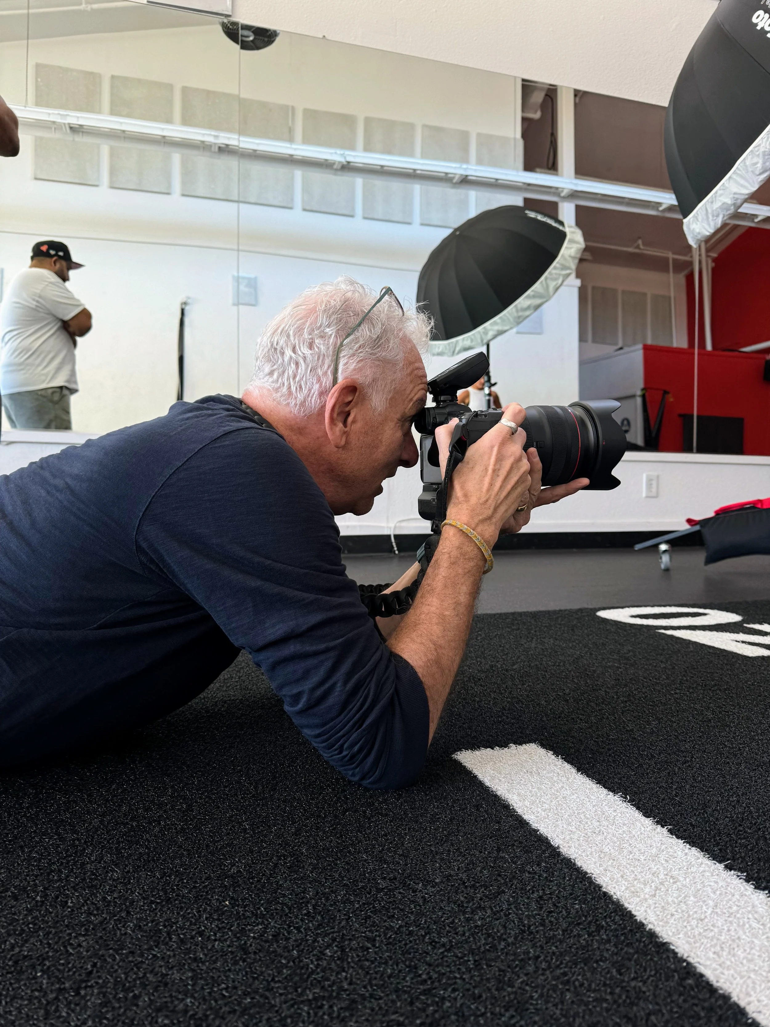A man with white hair lying on the floor and taking a photograph with a professional camera, in a studio with photography equipment and a mirror in the background.