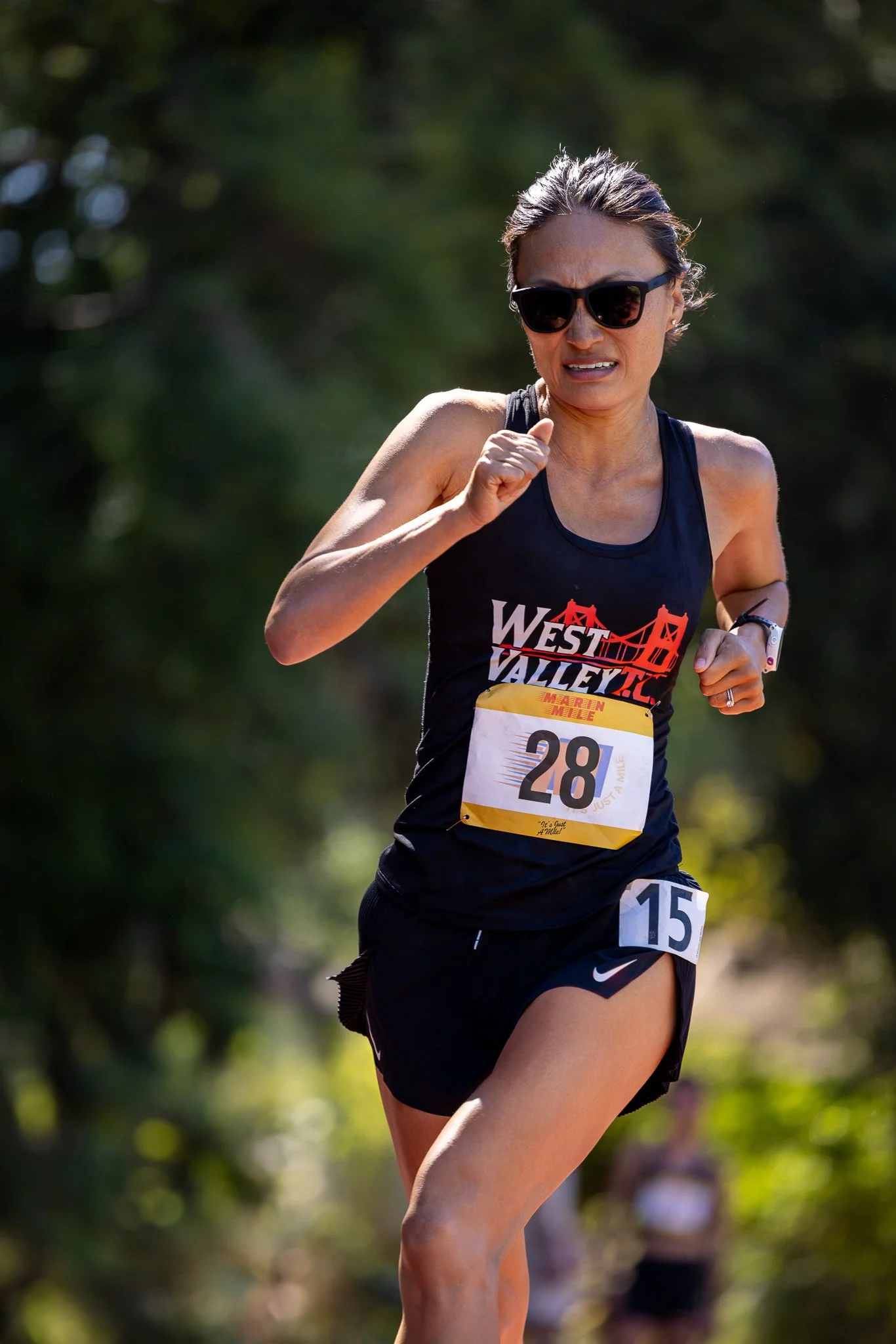 A woman running in a race, wearing sunglasses, a black tank top with 'West Valley' printed on it, and black shorts, with a bib number 28 and a race sponsor name Maren Mile, on a sunny day with trees in the background.