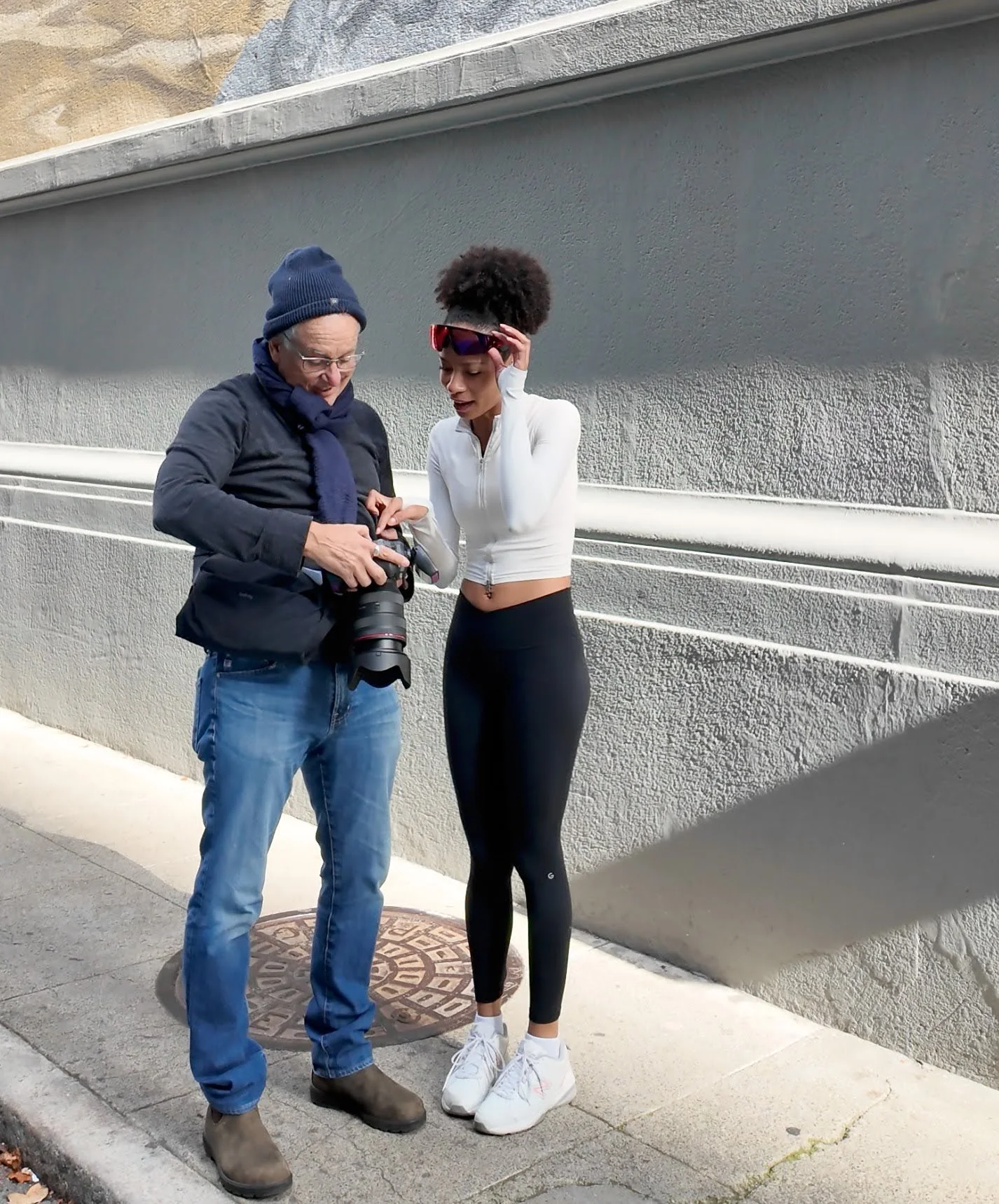 Two people, an older man with glasses and a beanie, and a young woman in athletic wear, looking at a camera together outdoor on a sidewalk near a grey wall.