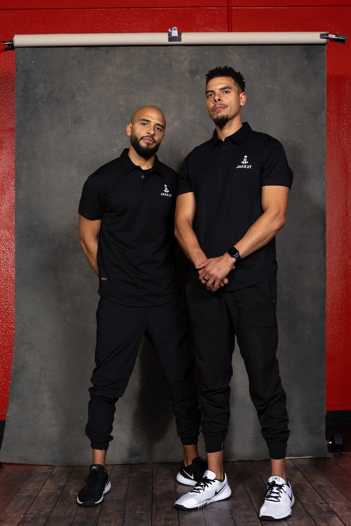 Two men standing side by side, wearing black polo shirts with a white logo and black joggers, posing in front of a gray backdrop in a photo studio.