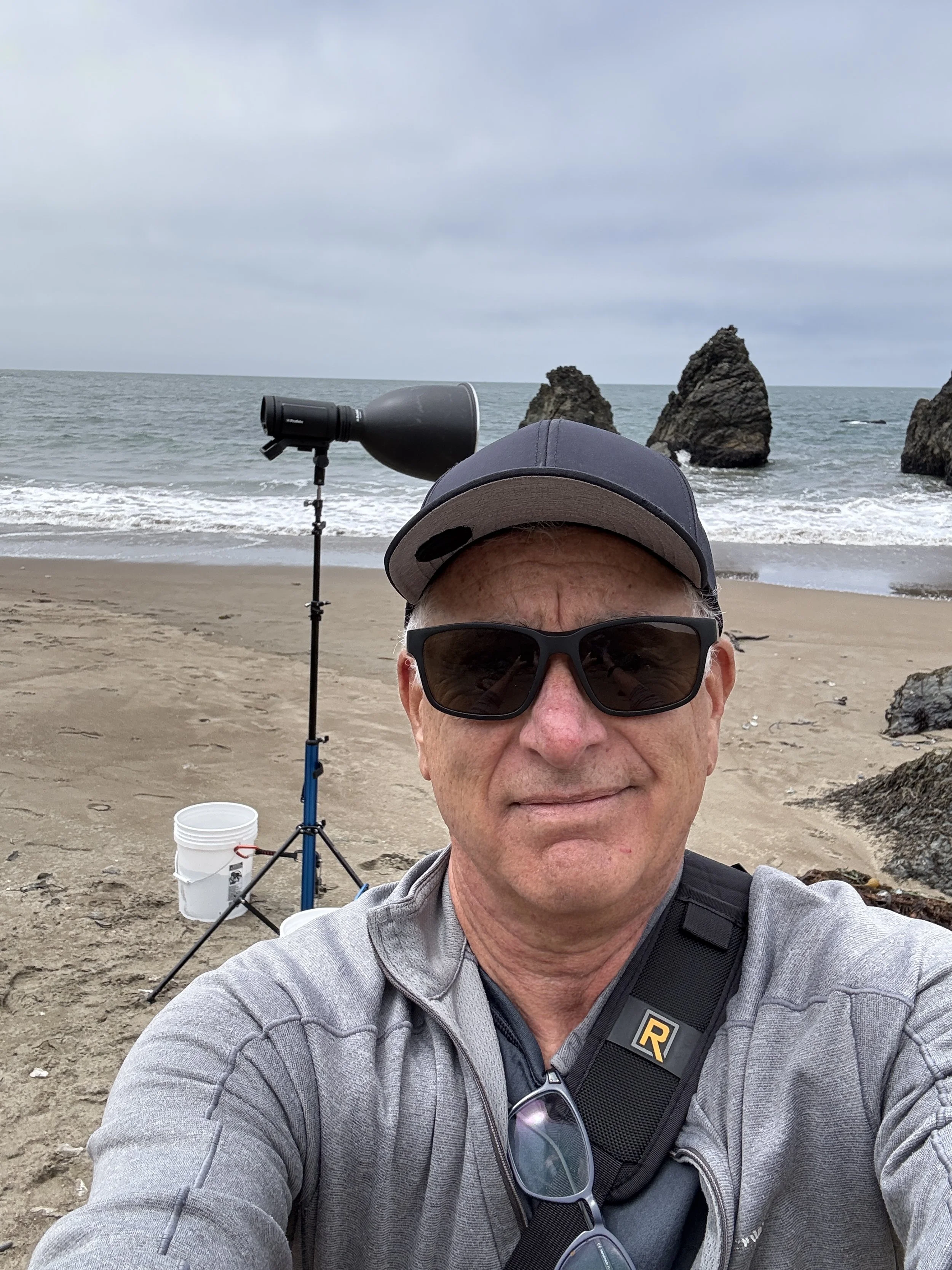 A man wearing sunglasses, a gray jacket, and a baseball cap takes a selfie on a beach with large rocks offshore. A spotting scope on a tripod and a bucket are also on the sand.