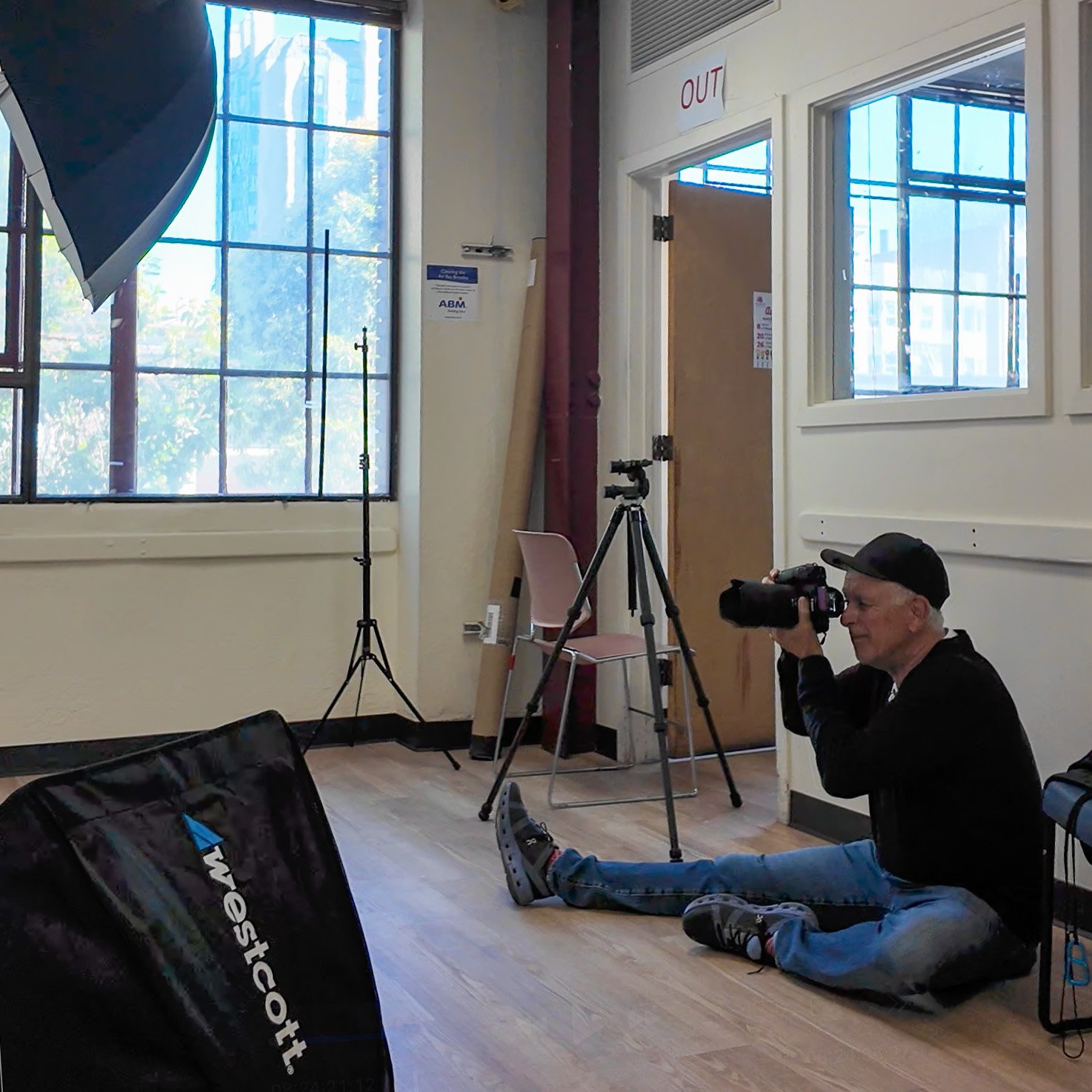 A man sitting on the floor uses a camera with a large lens, aiming it towards a window. The room has photography equipment including a tripod, a light stand, and a softbox labeled 'Westcott' on the floor. There is a pink chair, a door with an 'OUT' s