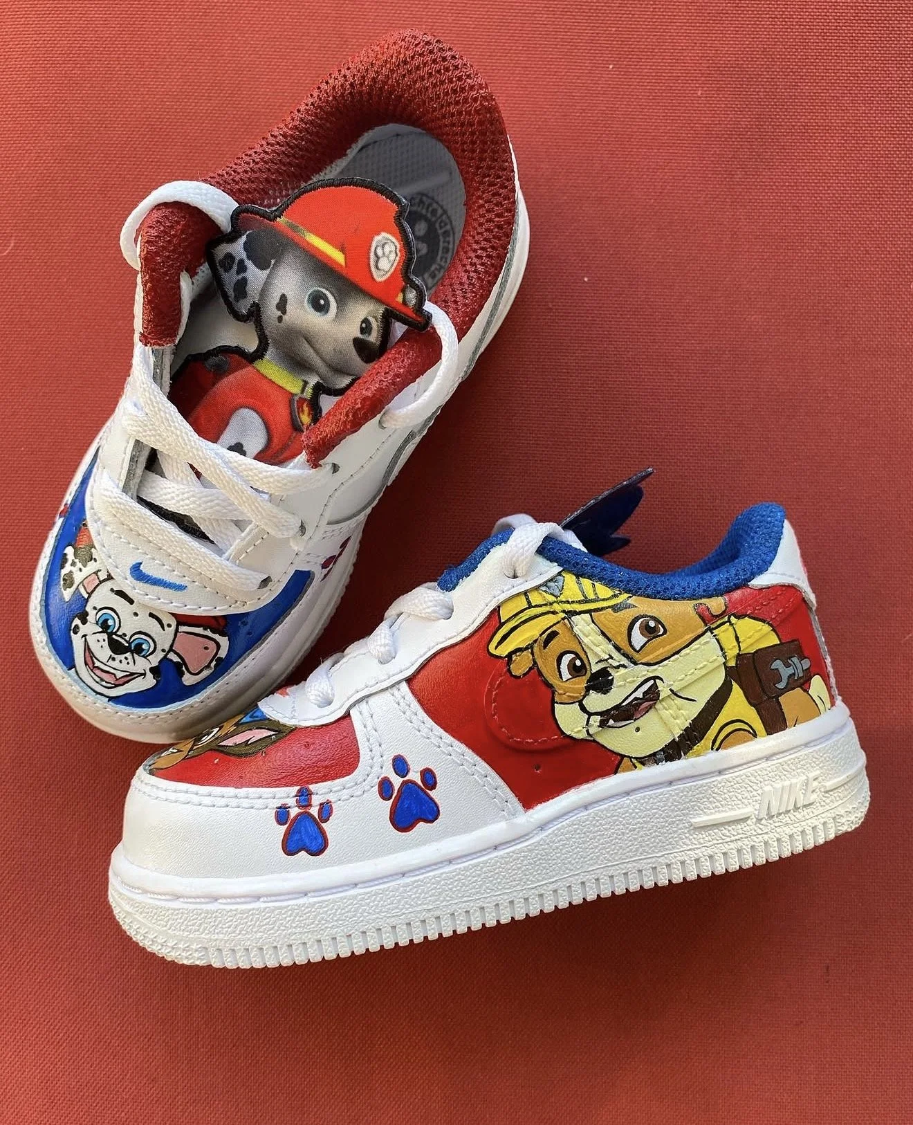 Child-sized white sneakers with red and blue accents, decorated with characters from a children's cartoon show, featuring dogs in firefighter and police costumes, on a red surface.