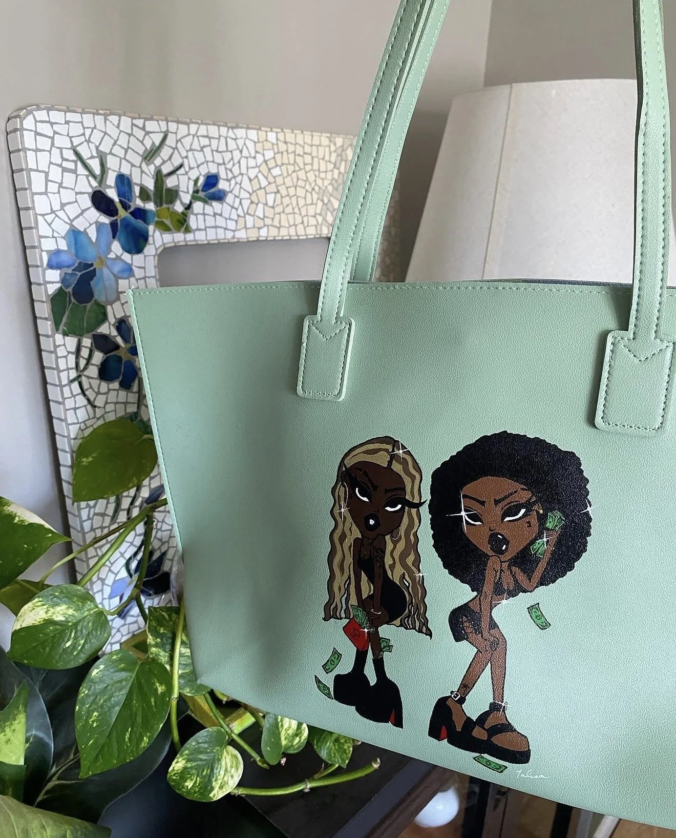 A mint green tote bag with cartoon illustrations of two women, one with long, blonde hair and the other with curly black hair, both holding dollar bills, placed near a mosaic mirror with blue flowers and green leaves in the background.