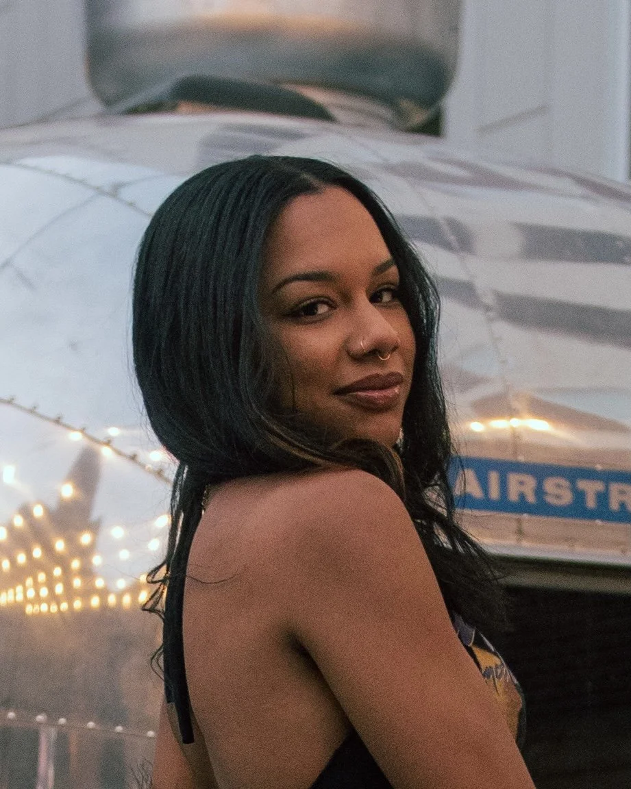 A woman with dark, shoulder-length hair, wearing a black top, stands in front of a small airplane decorated with string lights.