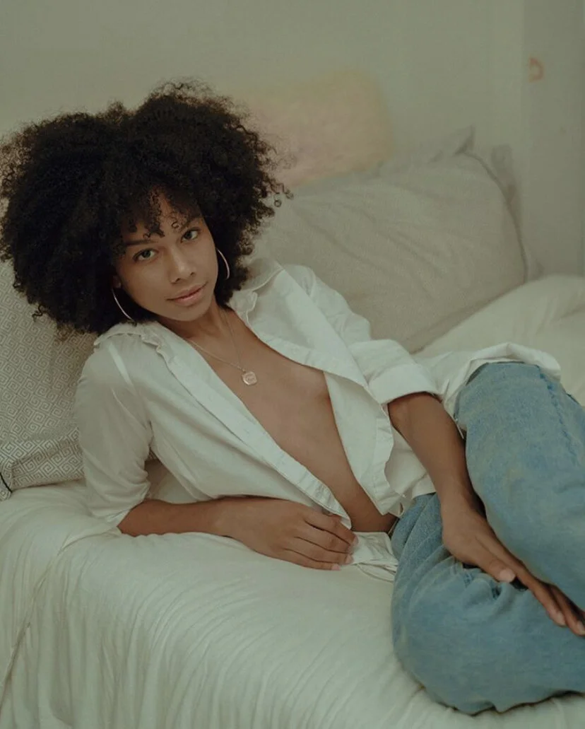 A woman with curly hair relaxing on a bed, wearing a partially unbuttoned white shirt and jeans.