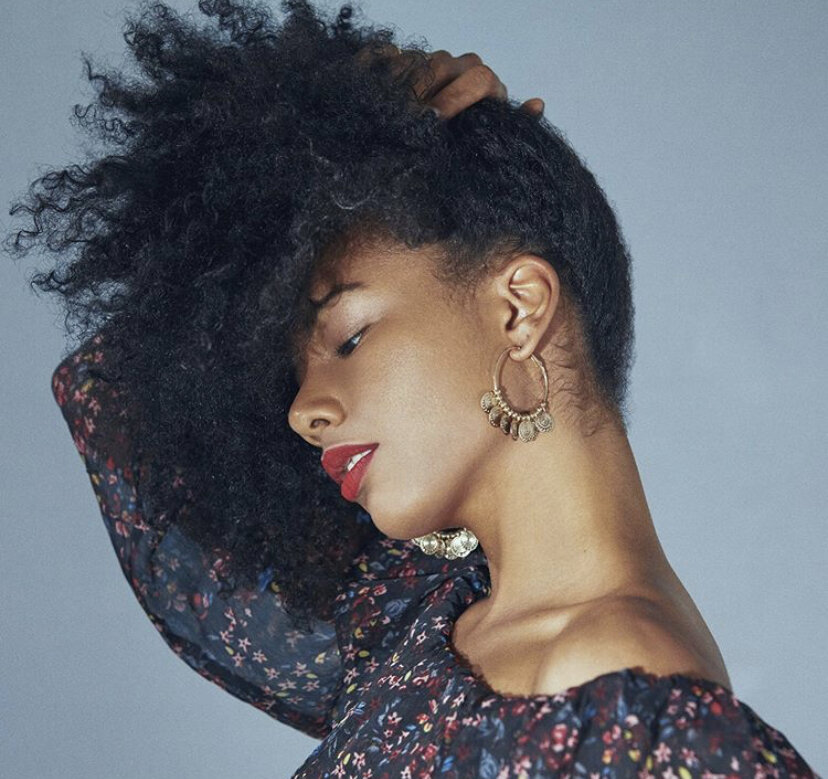 A woman with curly black hair, wearing red lipstick and gold earrings, posing with her hand on her head against a plain gray background.