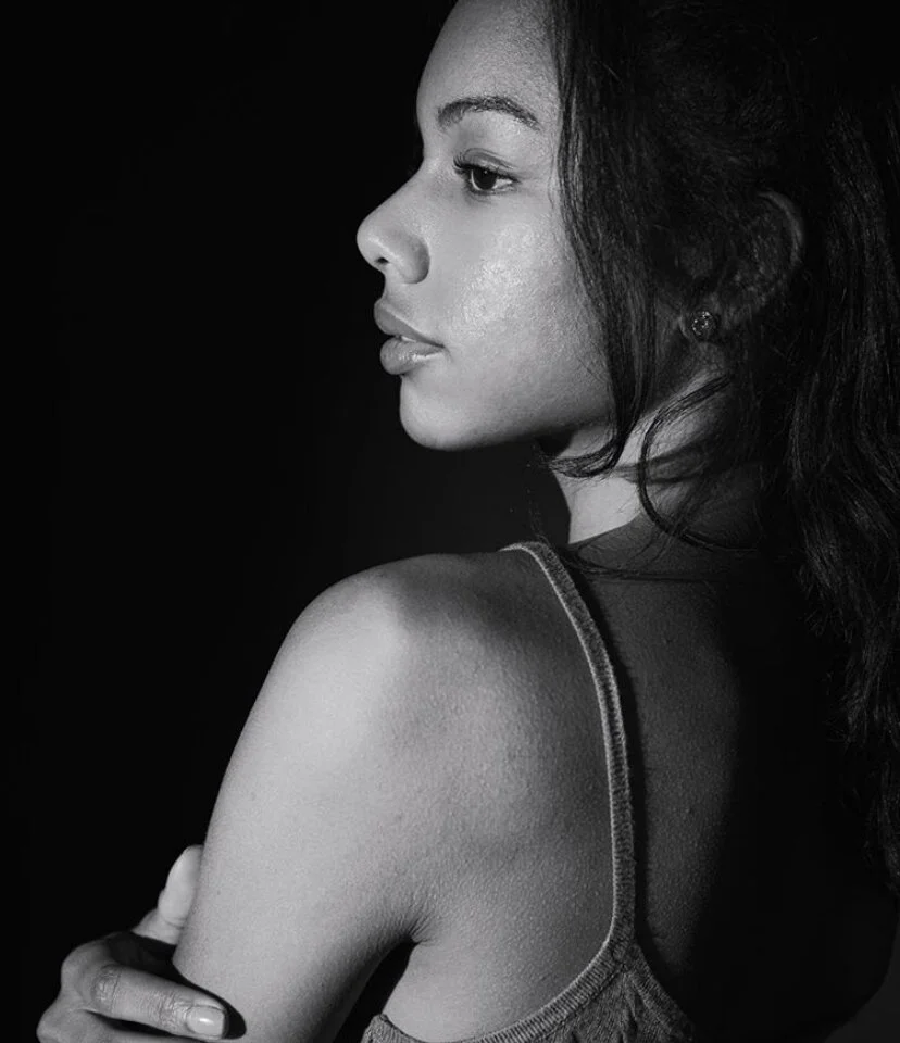 A woman with long dark hair looks to the side, wearing a sleeveless top, in a black and white portrait.
