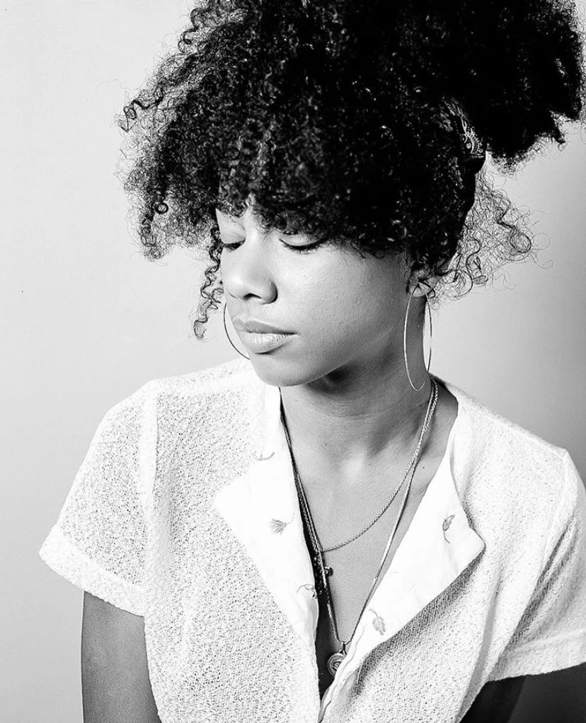 Black woman with curly hair wearing hoop earrings and layered necklaces.