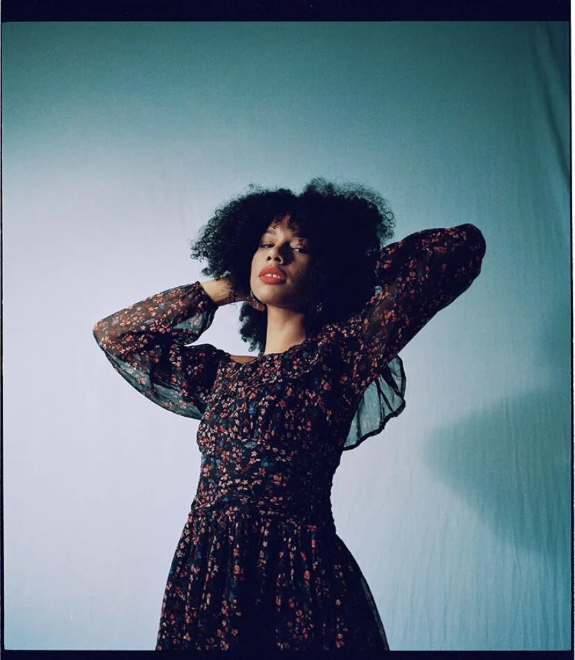 A woman with curly black hair posing with her hands behind her head, wearing a long-sleeved floral dress against a gradient teal and white background.