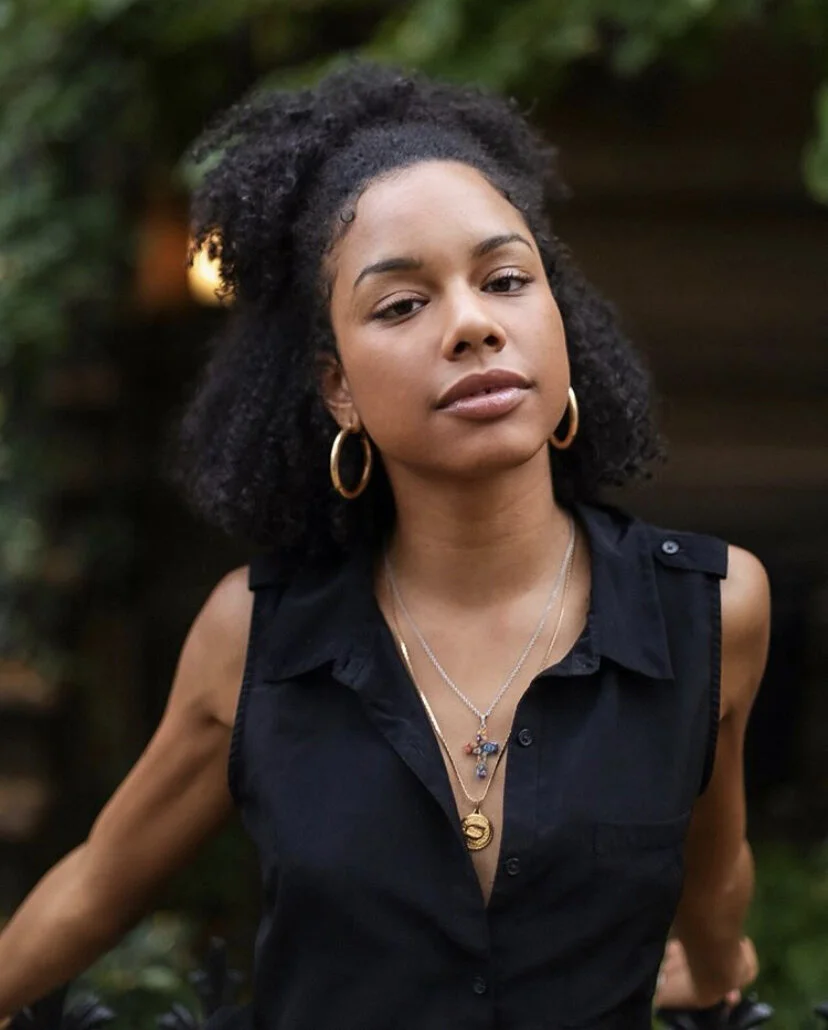 A young woman with curly black hair styled in a natural afro, wearing gold hoop earrings and layered necklaces, dressed in a sleeveless black top, standing outdoors with blurred greenery in the background.