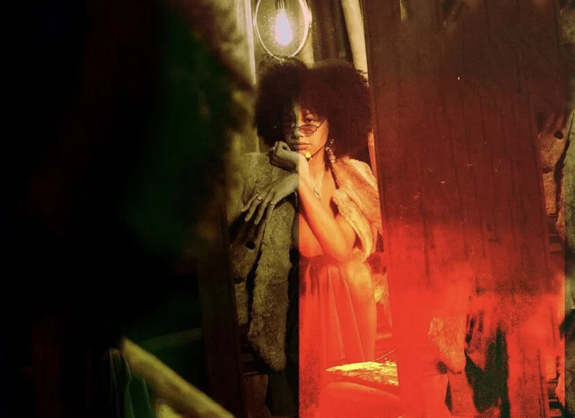 A woman with curly hair and glasses sitting in a dimly lit room with hanging light bulbs and wooden walls.