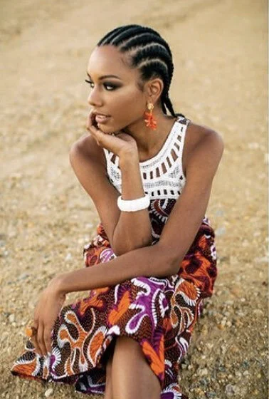 A woman with braided hair sitting on a dirt ground in a patterned skirt and white top, looking thoughtful.