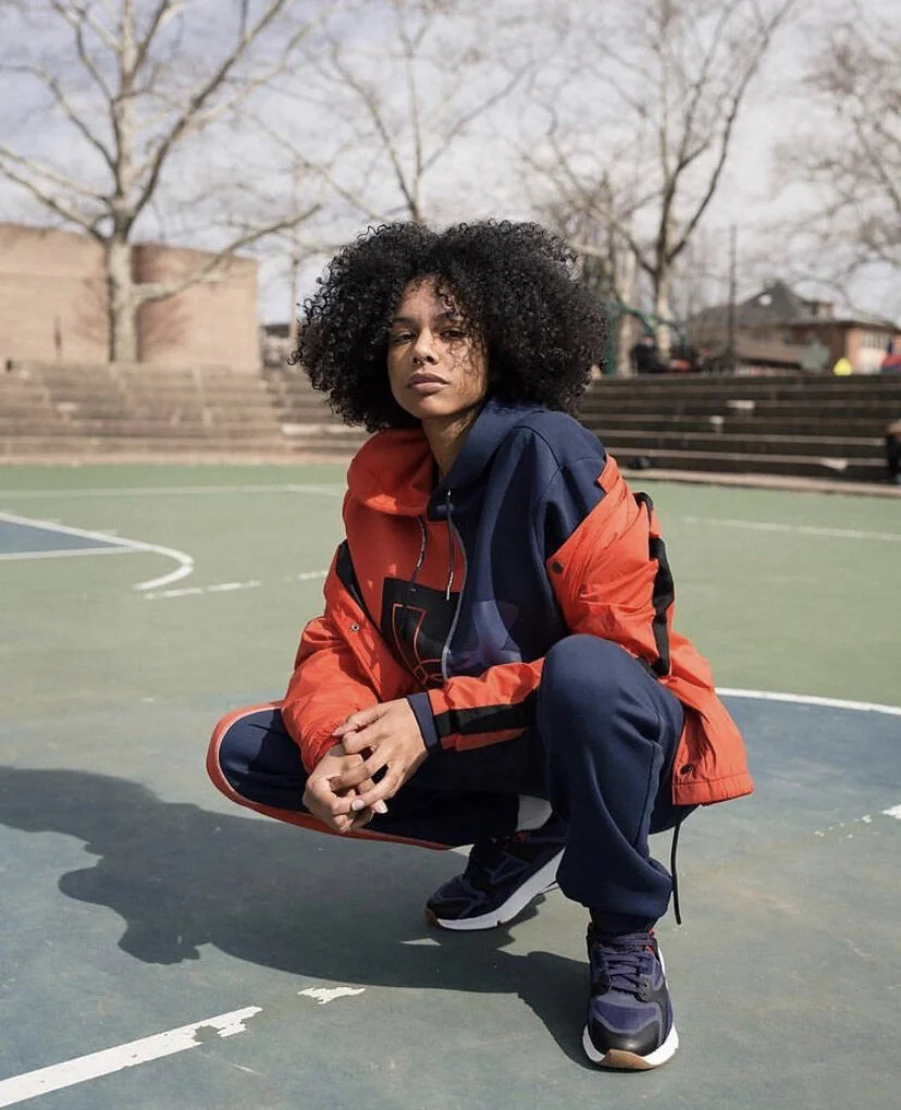 A woman with curly hair squatting on an outdoor basketball court, wearing a red and blue jacket and athletic shoes.