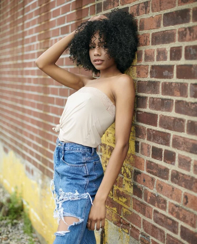A young woman with curly black hair leaning against a brick wall, looking at the camera, wearing a strapless beige top and ripped blue jeans.