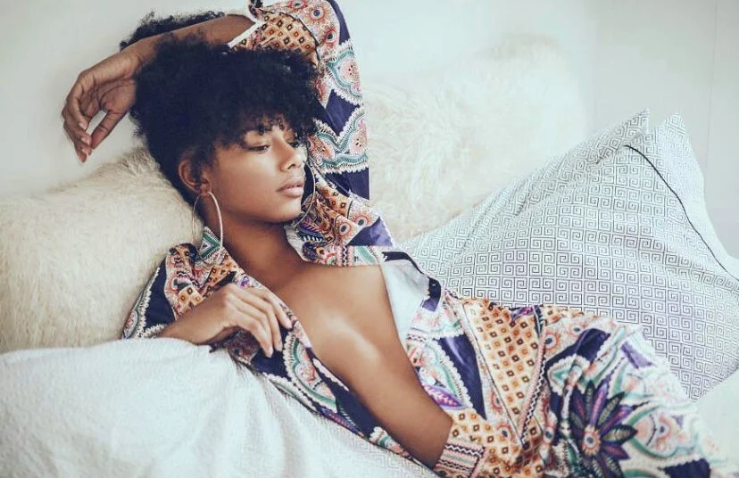 A woman with curly hair relaxing on a bed, wearing a colorful patterned shirt partially off, with one arm resting above her head and the other hand on her chest.