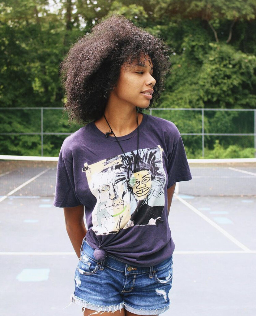 Young woman with curly hair standing on a tennis court, wearing a graphic t-shirt and denim shorts.