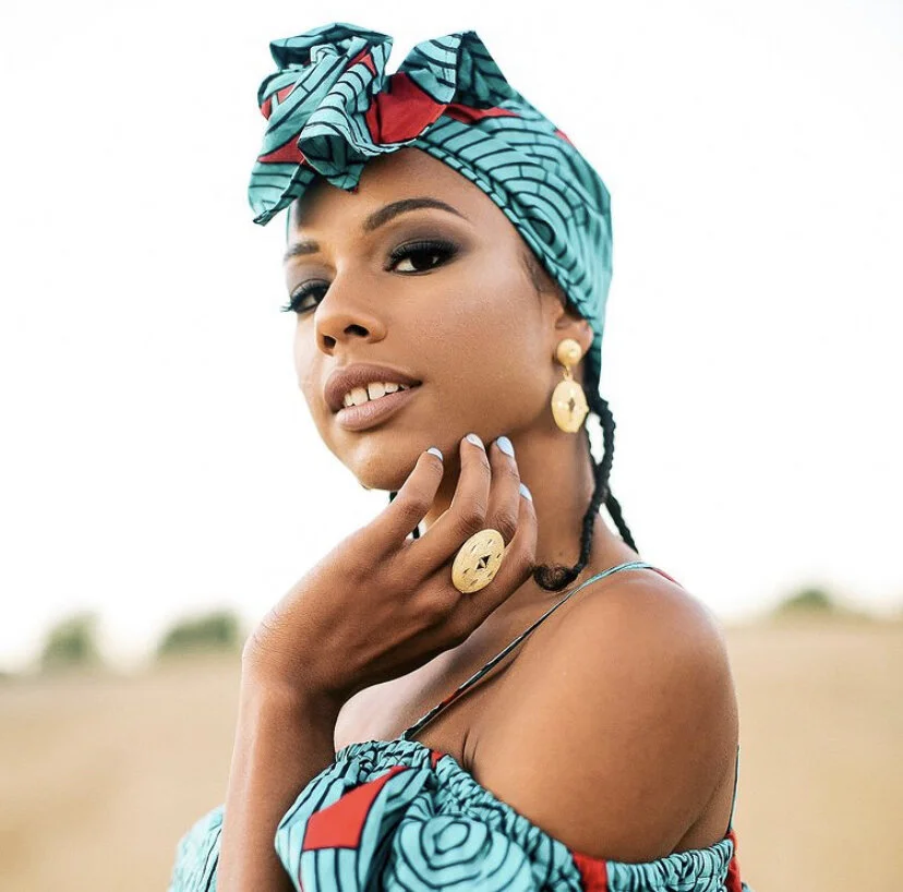 A woman wearing a teal and red patterned headwrap and matching off-shoulder dress, accessorized with gold jewelry, standing outdoors with a blurred landscape background.