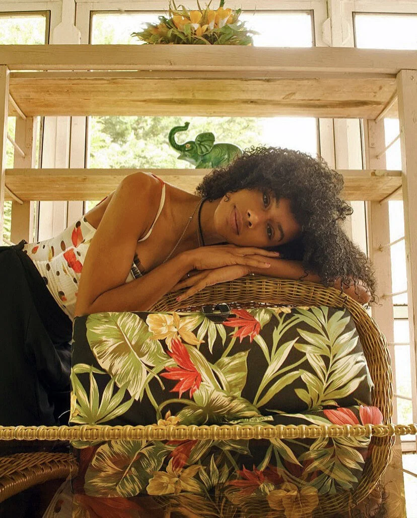 A woman with curly hair resting her head on her folded arms at a glass table with a floral cushion, with a background of wooden shelves, a decorative plant, and a window showing greenery outside.