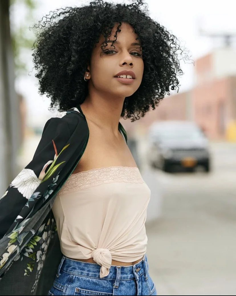 A young woman with curly black hair wearing a beige strapless top with lace detail, tied at the waist, and blue jeans, standing outdoors in an urban area with blurred buildings and a parked car in the background.