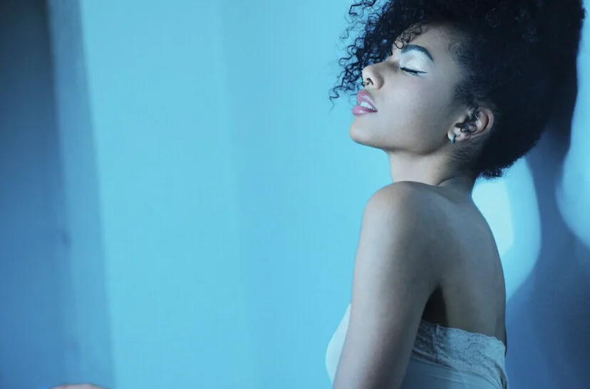 A woman with curly hair leaning against a blue wall with her eyes closed.