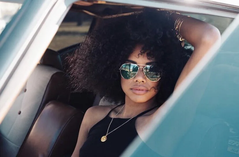 A woman with curly hair wearing sunglasses, a black sleeveless top, and gold jewelry sitting in a vintage car.