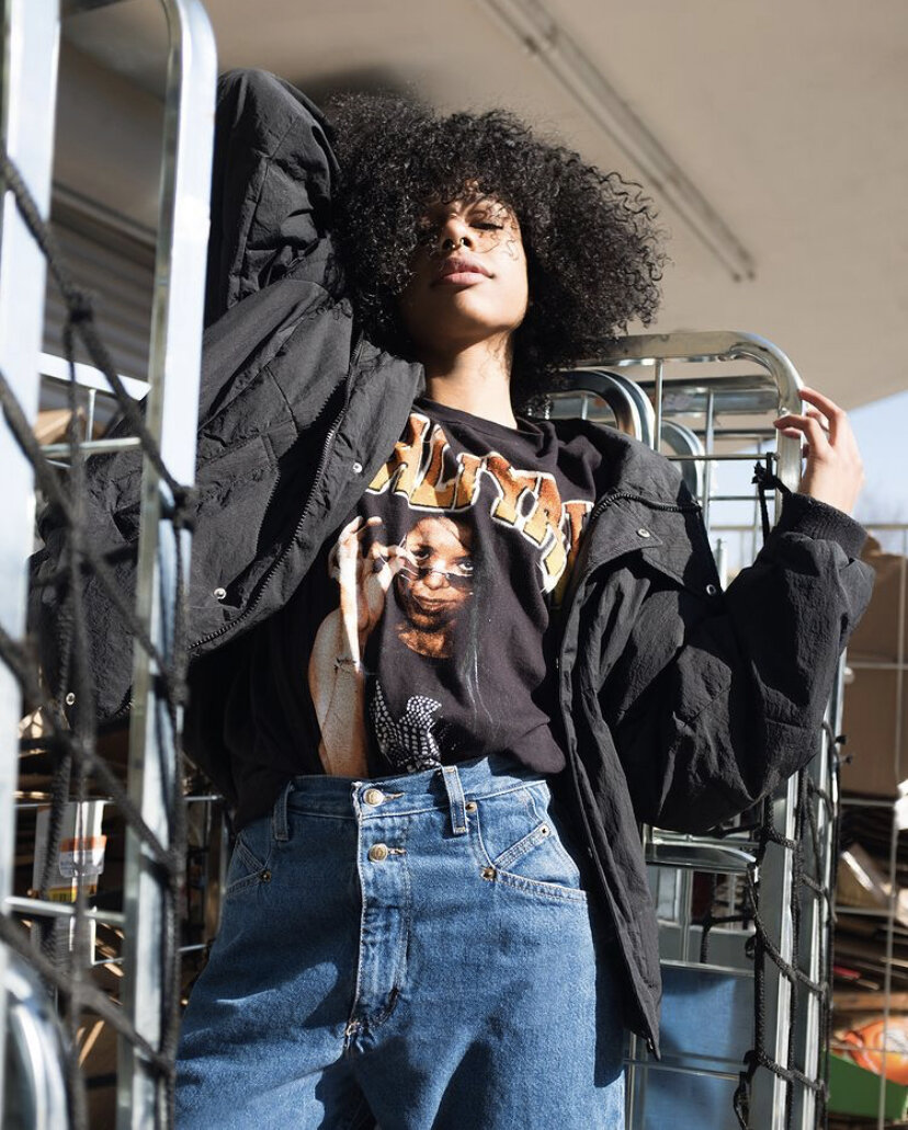 Young woman leaning against a metal cart or rack outdoors, wearing a black graphic t-shirt and an oversized black jacket, with curly hair and a confident expression.