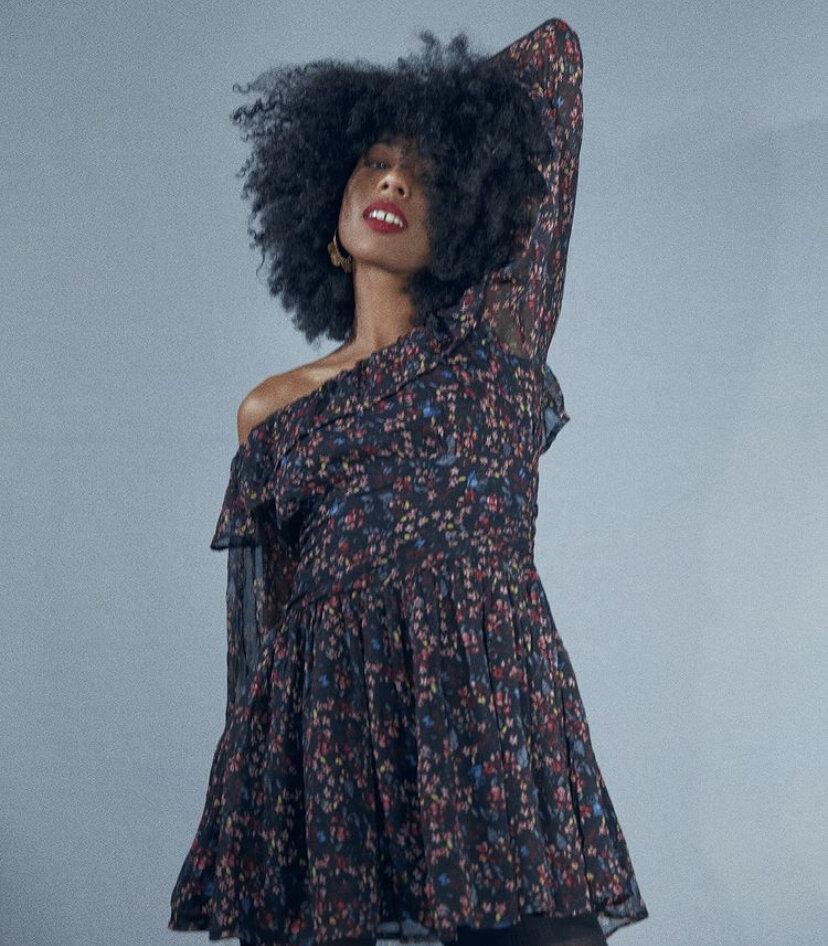 Woman with curly black hair wearing a floral off-shoulder dress, posing against a plain background.