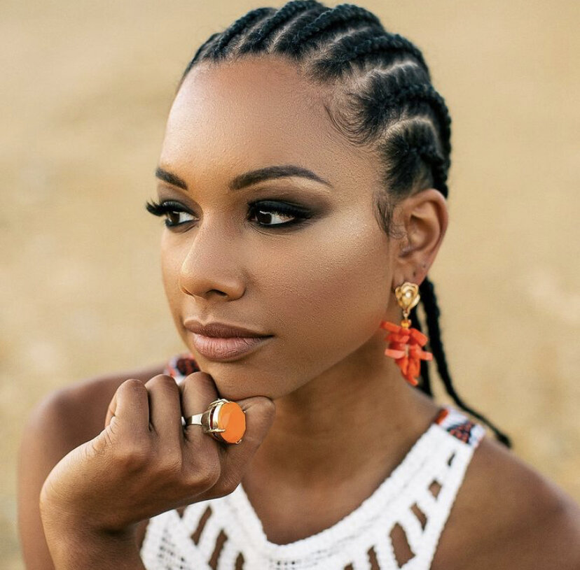 A woman with braided hair, wearing earrings and an orange ring, resting her chin on her hand outdoors.
