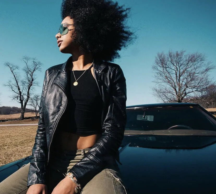 Young woman with curly hair wearing sunglasses, a black leather jacket, and a black crop top, sitting on a black sports car outdoors on a clear day with leafless trees in the background.