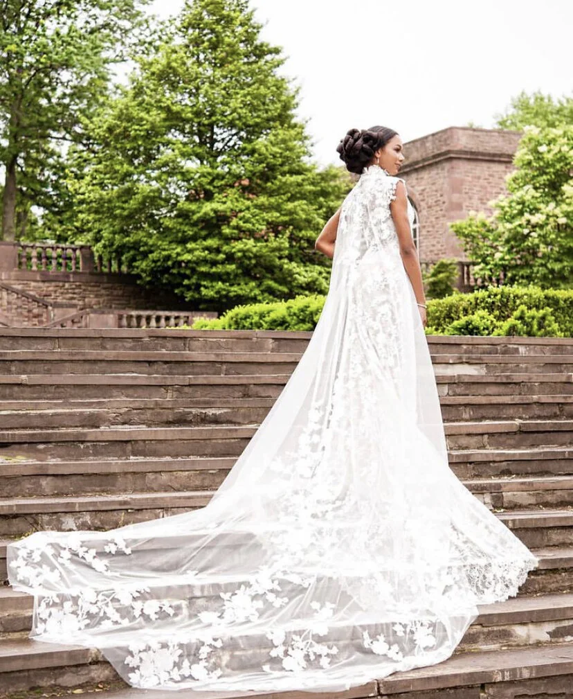 A woman in a white wedding dress with a long train standing outdoors on stone steps surrounded by green trees.