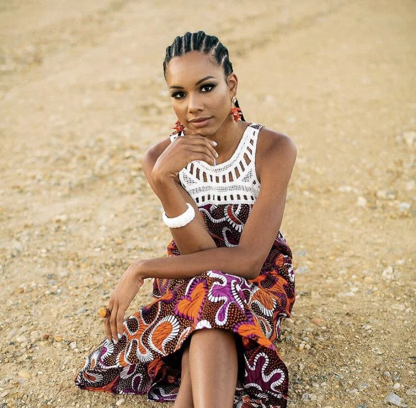 A woman sitting on a rocky ground, wearing a colorful patterned dress and earrings, with her hand resting on her chin.