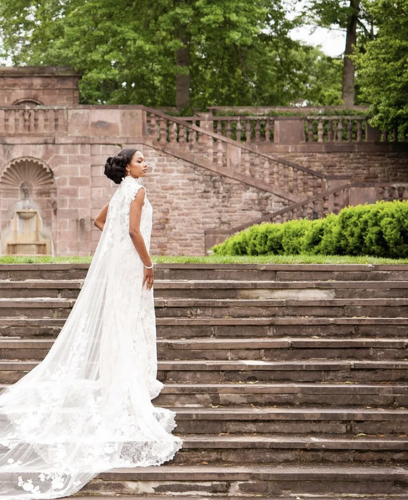 A bride wearing a white wedding dress with lace details standing on outdoor stone steps, with a historic stone wall and lush green trees in the background.