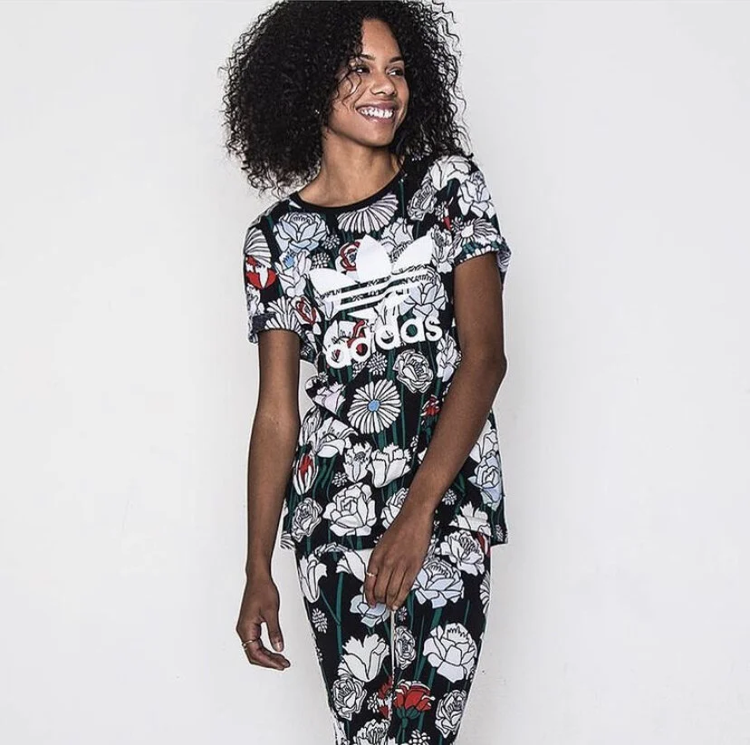 A woman with curly hair wearing a matching floral Adidas T-shirt and leggings, smiling and standing against a plain white background.