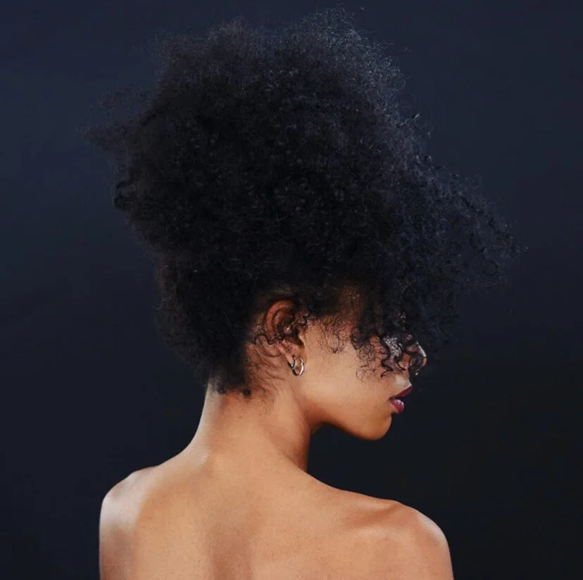 Side profile of a woman with curly black hair styled in an afro, wearing silver earrings, against a dark background.