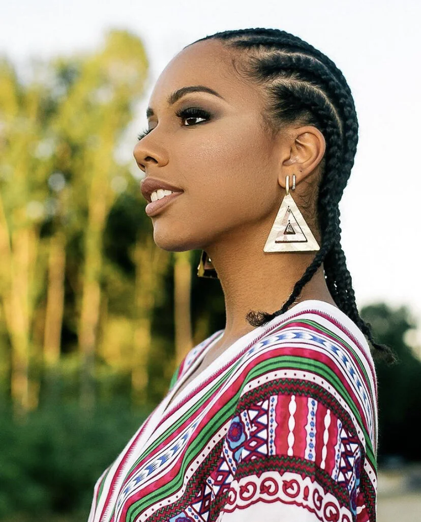 Profile of a woman with braided hair, wearing large geometric earrings and a colorful, patterned top, outdoors.