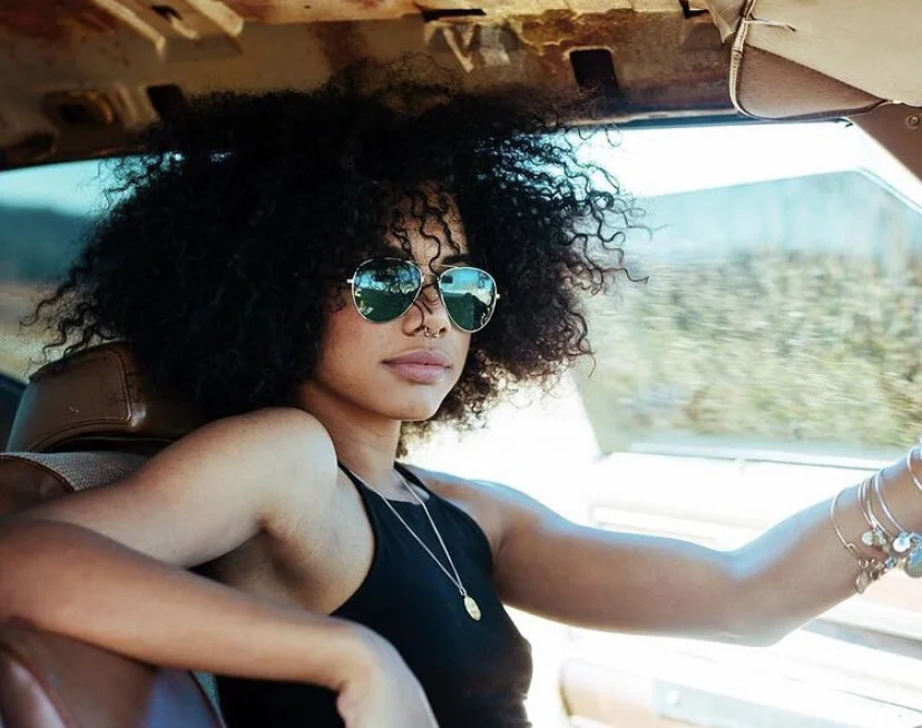 Woman with curly hair wearing sunglasses and jewelry driving a vehicle.